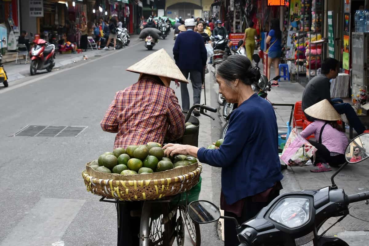 Marchandes ambulantes avec un panier d'agrumes sur un vélo dans une rue animée du vieux quartier de Hanoï, entre scooters et boutiques