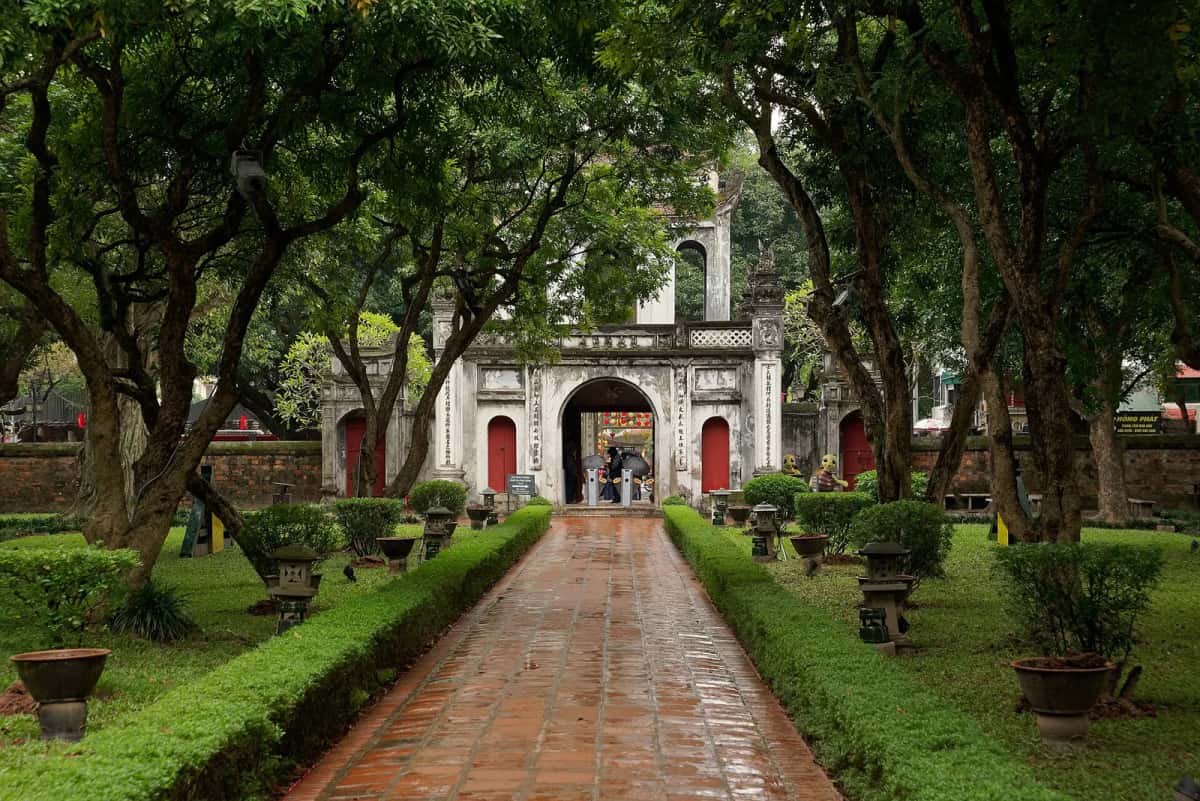 Allée dallée de brique menant à la porte principale du temple de la Littérature (Văn Miếu), entre des haies taillées et de grands arbres, à Hanoï