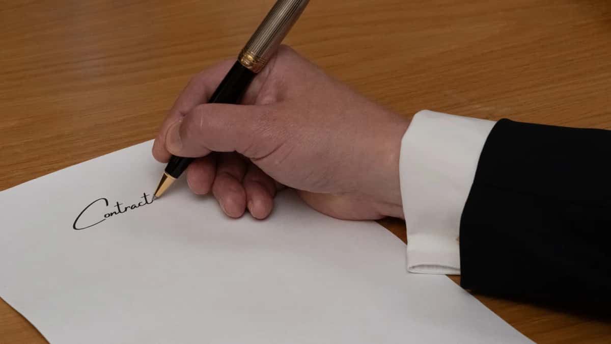 Main d'une personne en costume signant un document avec un stylo plume sur un bureau en bois