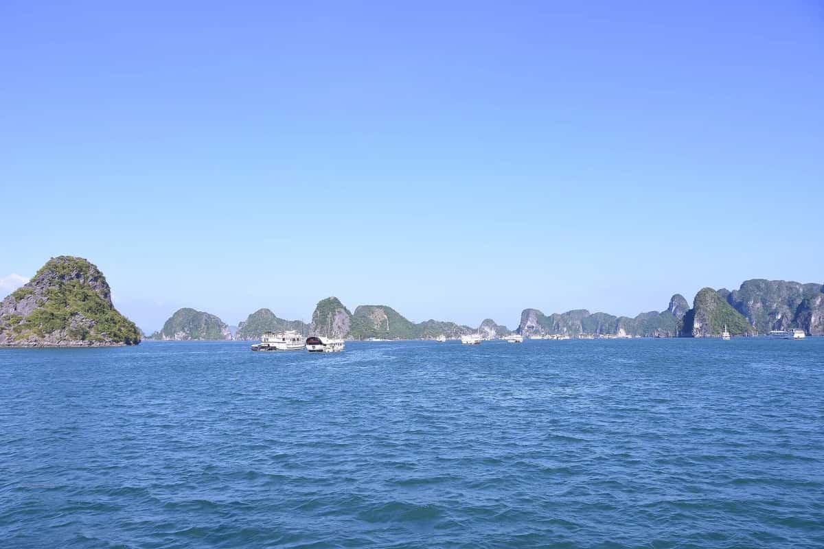 Îlots karstiques couverts de végétation et bateaux blancs sur une mer calme, sous un ciel bleu dégagé, dans la baie d'Halong