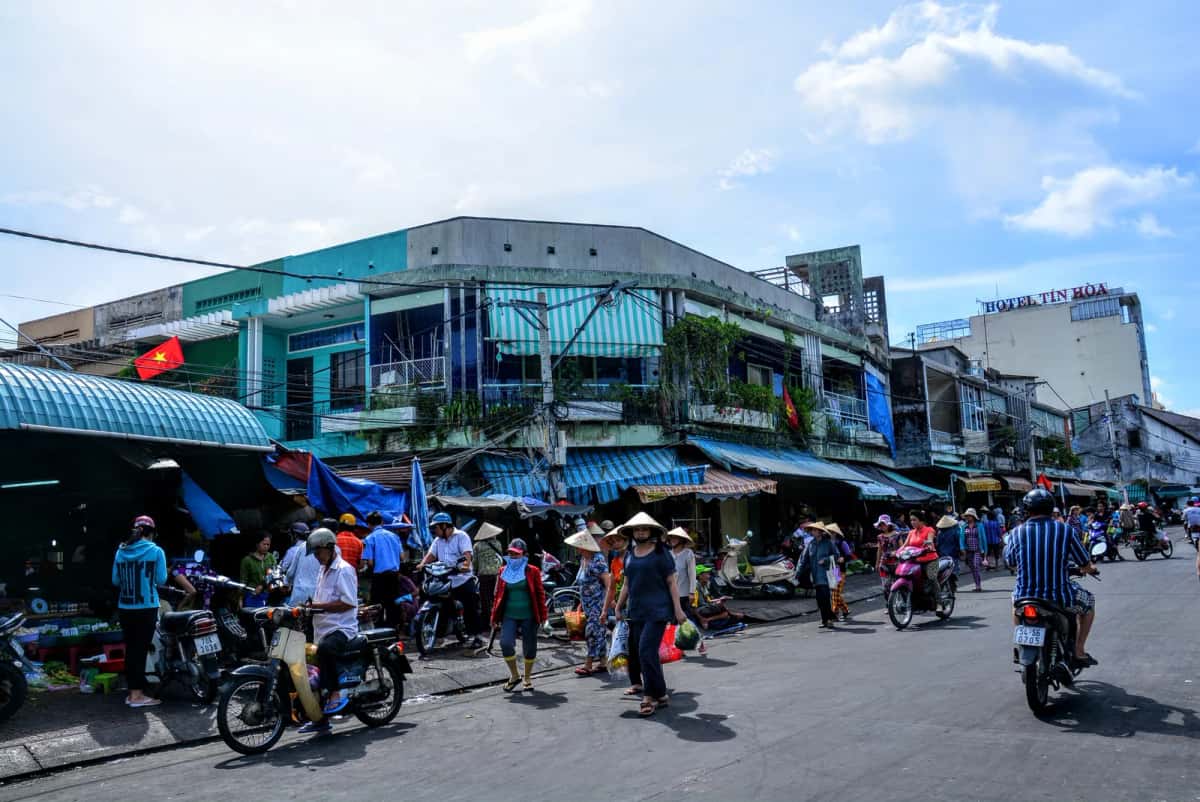 Carrefour animé devant un marché de quartier à Hô Chi Minh-Ville, avec des motos, des chapeaux coniques et des étals sous bâches