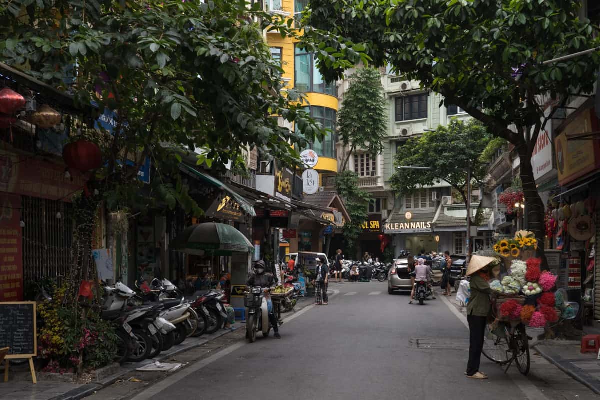 Rue arborée du Vieux Quartier de Hanoï avec des motos garées et une vendeuse de fleurs à vélo